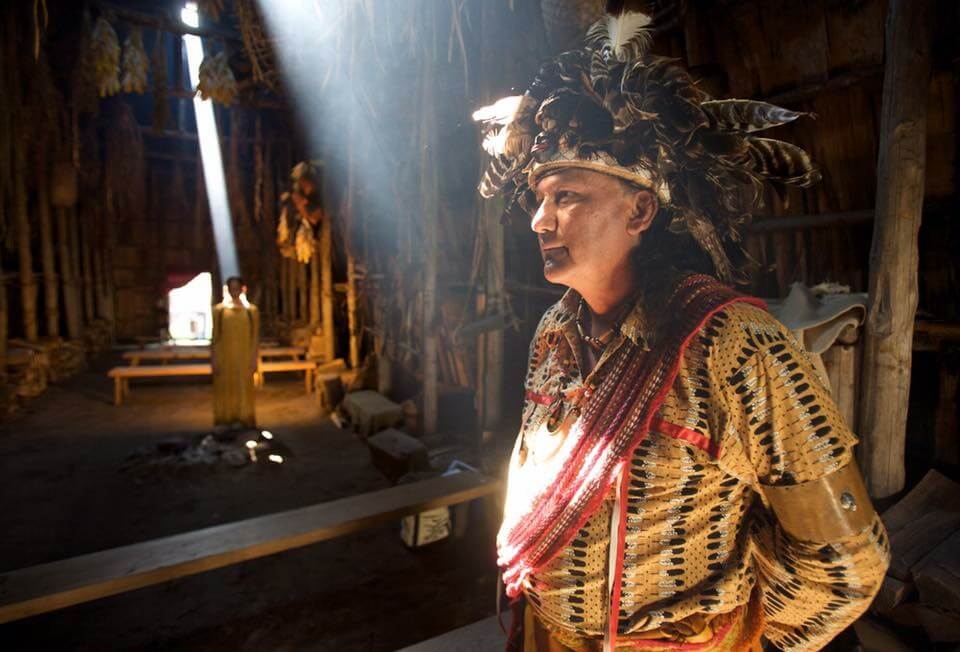 An Indigenous leader with a large headdress and metis sash, in the longhouse