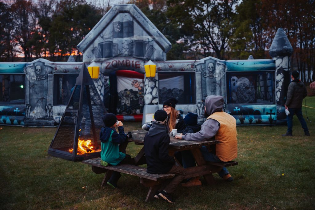 A family sitting at a picnic table and drinking hot chocolates outside the giant haunted house bouncy castle at Pumpkinferno