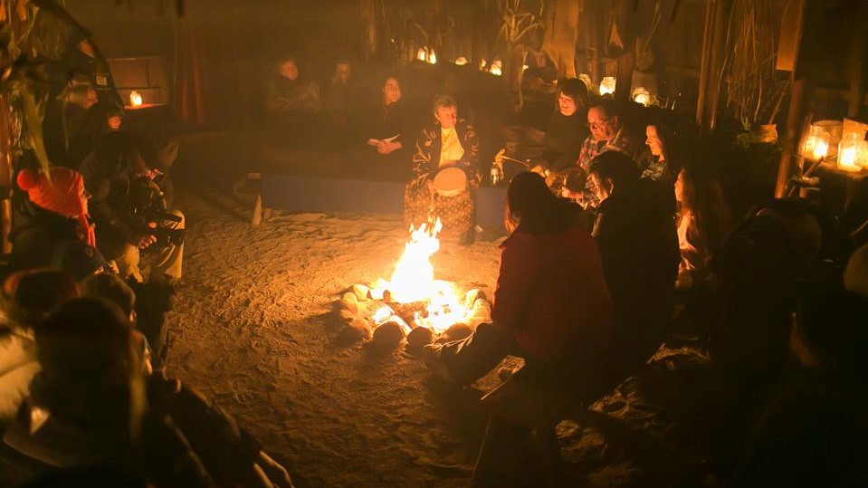 The inside of the longhouse, filled with guests and Indigenous elders sitting around a fire, surrounded by lit candles