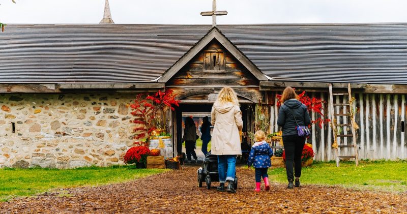 A family pushing a stroller and entering Sainte-Marie's main gate, which is decorated for fall with mums, sumac and gourds