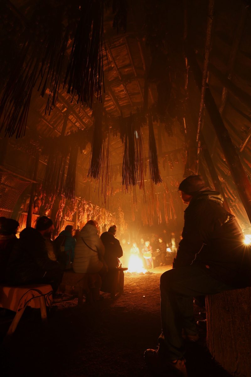 group of individuals sitting in front of a fire in the longhouse