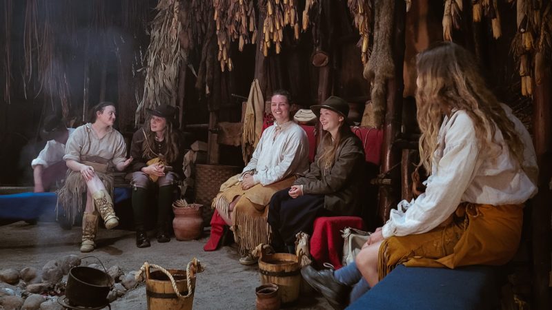 A group of costumed historical interpreters seated in the longhouse at Sainte-Marie